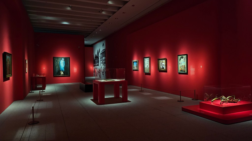 ‘Fantastic Visions: 100 Years of Surrealism from the National Galleries of Scotland’, installation view at Museum of Art Pudong. Photo: ArtsHub. A dimmed gallery space with red walls. Several paintings are hang and a sculpture is set atop a low plinth on the right hand side.