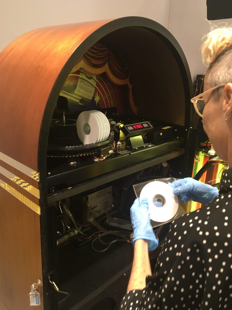 Lisa Mansfield working on the jukebox by artist Susan Hiller, ‘Die gedanken sind frei (Thoughts are free)’, 2012 for ‘What Does the Jukebox Dream Of?’ at AGNSW. Photo: Supplied. A person with a blonde pixie cut wearing glasses and a black shirt with white dots removing a small white CD from its plastic package. In front of them is the inner compartment of a jukebox.