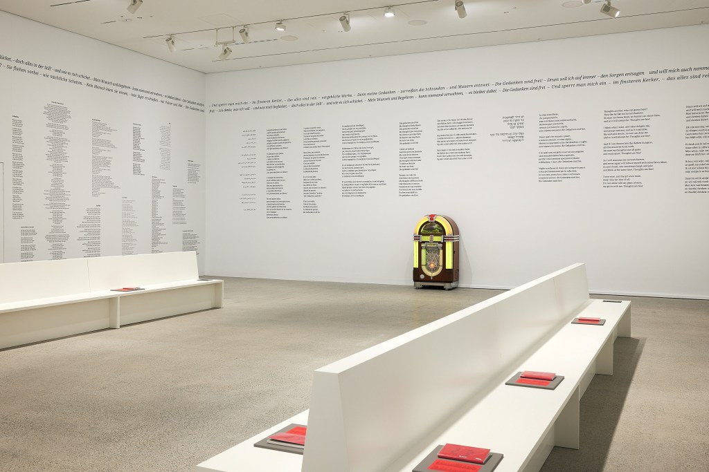‘What Does a Jukebox Dream Of?’, installation view at AGNSW. Photo: Supplied. A single jukebox with yellow neon lights stand inside a white-walled gallery space with concrete floor and two rows of white benches. On the walls are rows of (illegible) text, likely to do with the songs in the jukebox. On each bench are several booklets with a red cover.