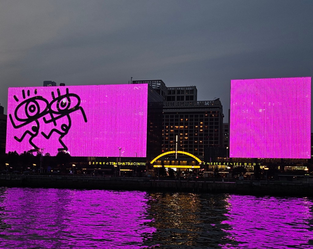 Large screens showing digital works by five local and international artists, seen from a harbour cruise. Image: Still from animation by Lousy. Photo: ArtsHub. Two large screens are projected with a fluorescent pink. On the left side are two line-figures mid-dancing with eyes as heads. The scene is viewed from a cruise on the water, with buildings in the background. 