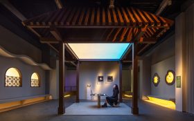 ‘The Hong Kong Jockey Club Series: Dwelling in Tranquility – Reinventing Traditional Gardens’, installation view at Hong Kong Palace Museum. Photo: Pak Chai. A woman sits in a spacious gallery space with a traditional Chinese architectural structure forming a shelter overhead. Windows of different patterns are illuminated on the surrounding walls.