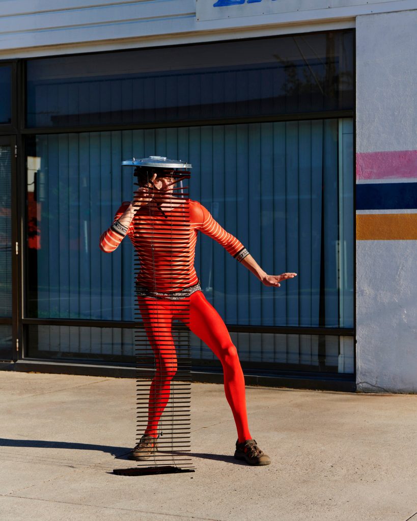 Jo Duck, ‘Venetian Sandwich’, as part of the series, ‘Razzle Dazzle’. Photo: Courtesy of the artist. A person in a red skin-tight suit peering out of some blinds that is attached to a metal tin lid on his head as a disguise. 