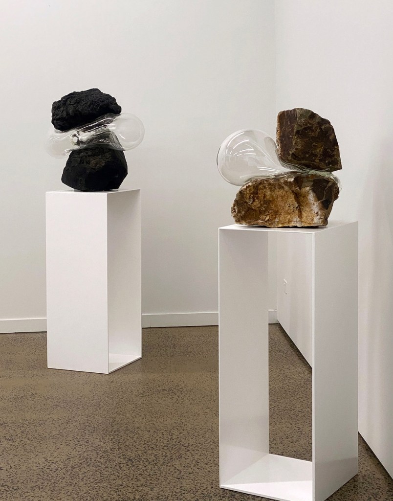 Rocks holding up. Simone Slee. image is four pieces of rock on two plinths in a gallery. Each pair are on top of each other with a bulbous glass artefact squeezing out from between them.