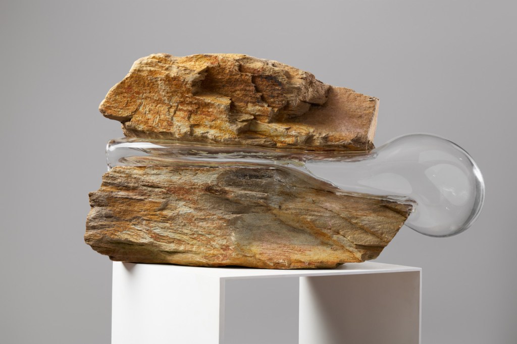 Rocks holding up. Simone Slee. image is two pieces of rock on a plinth. They are on top of each other with a bulbous glass artefact squeezing out from between them.