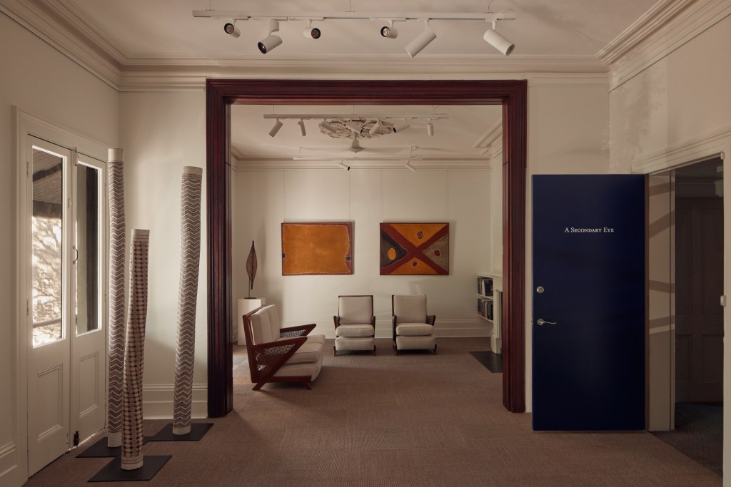 Installation view, A Secondary Eye Gallery. Featuring artworks L-R: Three Larraktij poles by Naruwapu Wunungmurra, a broad shield by an Unknown Wemba Wemba Artist, 'Lake Argyle' by Rover Thomas and 'Desert Meeting Place' by Rover Thomas. Photo: Supplied. A beige coloured gallery space during daytime. It appears to be housed in an apartment, with 'A Secondary Eye' letters on a black door half open.