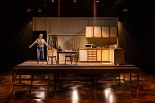 Sydney Theatre Company. Into the Shimmering World. Image is a stage set of a farmhouse kitchen, set upon a platform with stairs up to it. Three of the four walls are missing and a man in farmer's clothes stands on the left of the platform with arms outstretched.