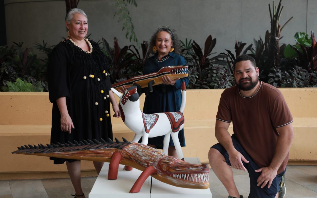 L-R: Fashion designer Irene Robinson, CIAF Artistic Director Francoise Lane, and CIAF curatorial associate Teho Ropeyarn celebrate the program launch of CIAF's 15th anniversary season. Assorted artworks by the artists from Wik & Kugu Art Centre. Photo: Supplied. Robinson is a woman with short grey hair wearing a black dress with a large necklace that has pompoms attached to it. Lane is a woman with curly grey hair, a warm smile, holding a carved crocodile artwork. Ropeyarn is a man with dark brown hair and a trimmed beard, kneeling beside two more wooden carved artworks, including another crocodile and a dog. The three of them are posed in a light-filled, outdoor space.