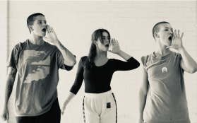 a black and white shot of three dancers (outside ones with shaved heads, middle with long hair and a fringe) in casual clothes standing in a row looking off to the right and with their left hands open next to their mouths as if they're shouting 'ahoy'.