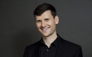 A headshot of a smiling white man in a black shirt holding a trombone in front of him.