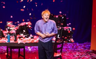 Josh Thomas. Let's Tidy Up. Image is a young man on stage in a blue shirt and black pants, surrounded by bits of furniture and objects and with confetti falling from the ceiling all around him.