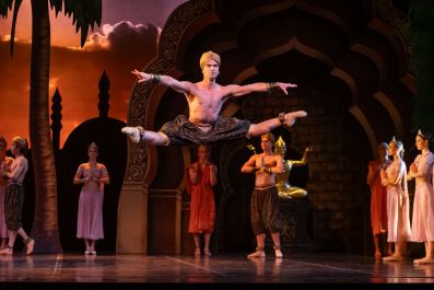 A bare-chested male ballet dancer dressed in a turban and long pants, photographed in mid air performing a leaping side split move.