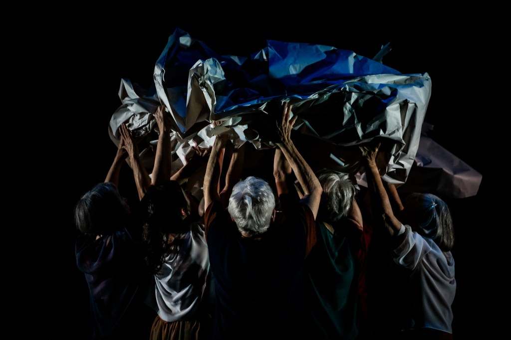 A body wrapped in paper is carried aloft by five women who have their backs to the viewer.