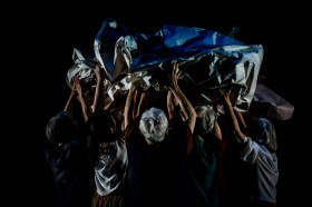 A body wrapped in paper is carried aloft by five women who have their backs to the viewer.