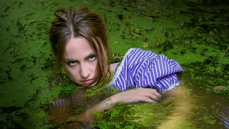MICF 2024 Belinda Anderson-Hunt. Image is a young woman in a purple and white striped shirt, mostly submerged in a pond that is covered in green slime/moss.