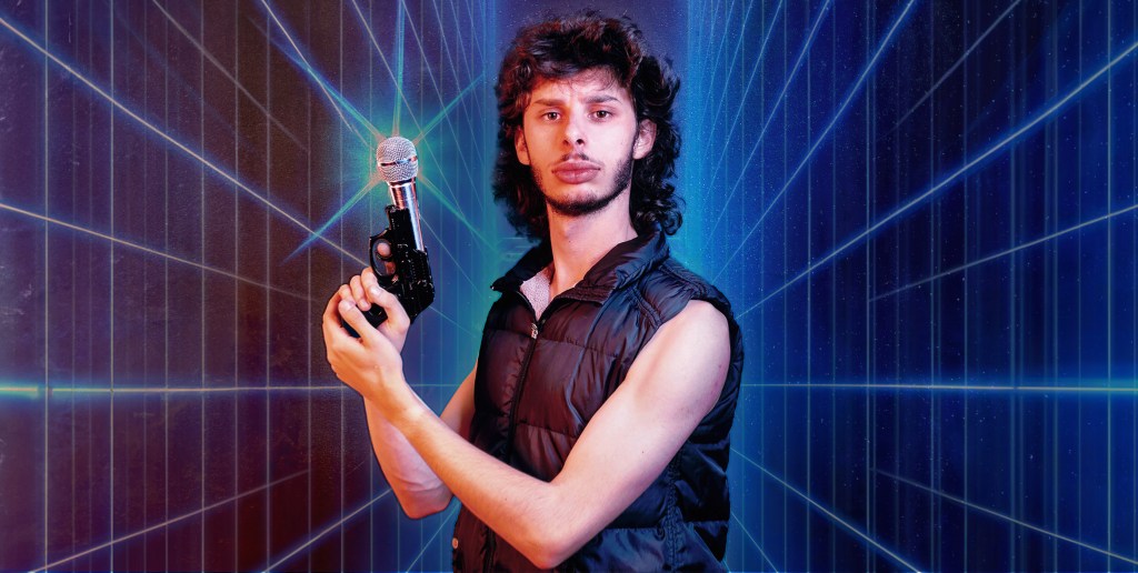 MICF 2024 Con Coutis. Image is a futuristic backdrop with squares travelling away as if lining a tunnel. In the middle is a young man of European appearance with a dark wavy mullet, scraggly beard and confronting expression. He is holding a gun with a mic in the end of it.