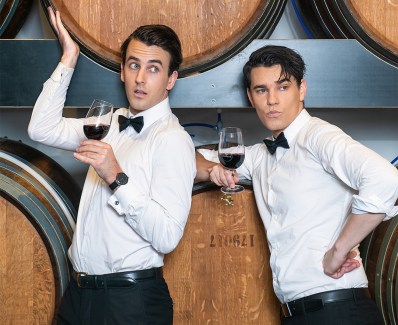 Two young white men in black pants, white shirts and black bowties, stand in front of large liquor barrels, brandishing glasses of red wine and looking off camera to the right. In Pour Taste: A Comedy Wine Tasting Experience. Sweeney Preston and Ethan Cavanagh.