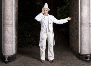 Julia Masli. ha ha ha ha ha ha ha. Image is a woman dressed in traditional white clown costume with a conical hat. She is standing between two pillars. On her white suit is written the word 'ha' several times.