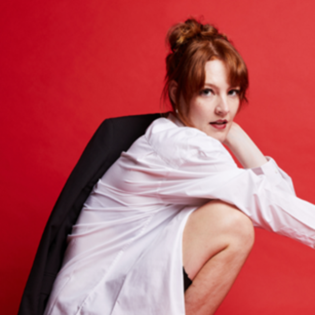 MICF 2024 Lou Wall, Image is a red background to a young red headed woman with hair in a scrunched bun wearing a white shirt, with a black jacket draped over her shoulder, squatting to the right and facing the camera.