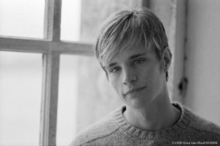 Matthew Shepherd. Image is a black and white head and shoulders shot of a young blond man wearing a jumper and standing in a room in front of a window. His head is tilted to the left.