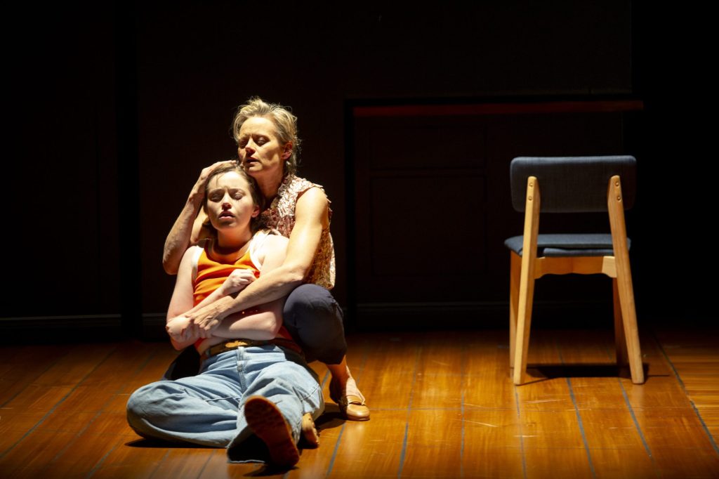 A bare stage aside from a single chair. To the left of it is a white teenaged girl looking anguished on the floor. An older white woman is behind her holding her and attempting to console her.