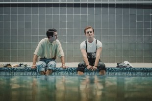 Streaming. Two young men sit next to a pool with their trousers rolled up and their legs dangling in the water.