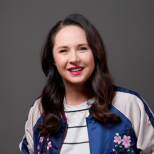 Mel Buttle. Image is a head and shoulders shot of a smiling woman with a side parting and long wavy hair wearing red lipstick and a stripey T shirt under a blue and white jacket with a floral pattern on the chest.