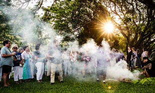 NT Writers Festival. Image is a group of people standing outside with the sun coming through the trees and observing a smoking ceremony.
