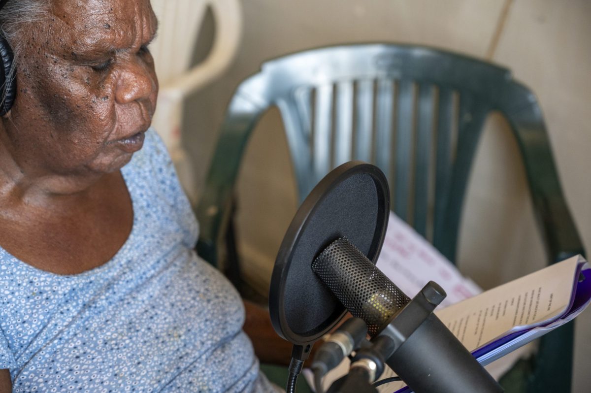 Australian country music has a complex history. Image is an older Aboriginal woman sitting in front of a microphone. A green plastic chair is behind her.