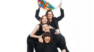 Four singers in black line up behind each other, first person crouching, so that the heads of each show above the person in front. The man at the back holds up a large perspex bowl full of coloured balls.