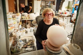 An elderly white female artist wearing bold red rimmed glasses in her ceramics studio.