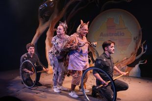 Possum Magic. Two actors in khaki style uniforms turn axles with a wheel on one side, while between them two white female actors are dressed as possums and are being transported. Behind the actors is a tree and a map of Australia on the back wall.