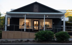 The front facade of REDSEA Gallery in Margaret River, housed inside a century-old building. Photo: Supplied. A black shed-like building taken from the outside. Leading up into the space is a long ramp with shrubs at the front. Inside the space are paintings showing through the gallery windows on both sides.