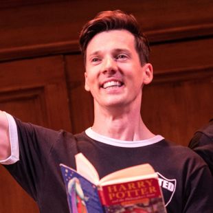 A young white smiling white man holds up one arm and in the other holds a Harry Potter book.