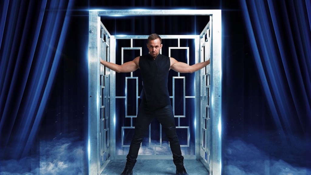 Magician dressed in black standing theatrically on stage with blue curtains. Anthony Street.