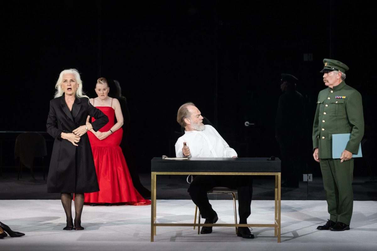 Four actors on a stage. On the left a white haired white woman in black, behind her a woman in a long red dress, seated at a desk is a middle aged white man with a beard, and on the right is a man in military uniform. The President.
