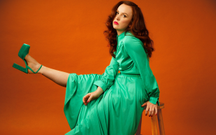 MICF 2024. Catherine Bohart, Again With Feelings. Image is a woman sitting on a chair, with her body facing the left side. Her right leg is kicked up in the air and she has a doleful expression on her face. She is against an orange background, has long wavy red hair and is wearing a green dress and high heeled shoes.