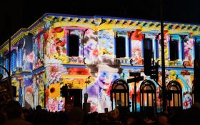 White Night returns to Ballarat. Photo: Supplied. Floral projections cast onto a building during the night, featuring vintage portraits that have flowers as eyes. A crowd gathers outside.