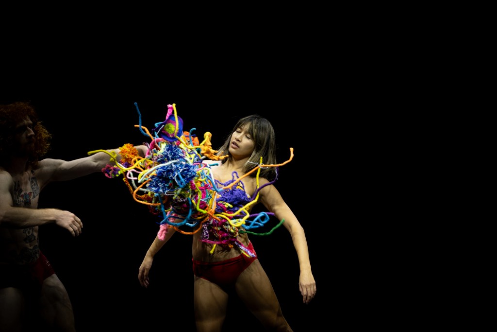 A young woman dancer has a mass of coloured fibres and fabrics hitting her in the chest.