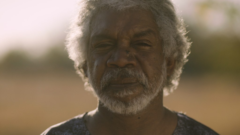 We Were Lost in Our Own Country. Image is a headshot of a middle aged Aboriginal man with a short grey beard and moustache, squinting his eyes at the camera.