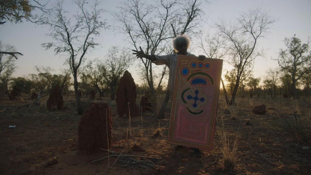 We Were Lost in Our Country. Image is an Aboriginal man on Country holding a big piece of card with abstract imagery on it and pointing to the right.