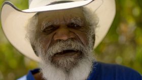 We Were Lost in Our Own Country. Image is a headshot of an elderly Aboriginal man with a white beard and moustache, wearing a white cowboy style hat.