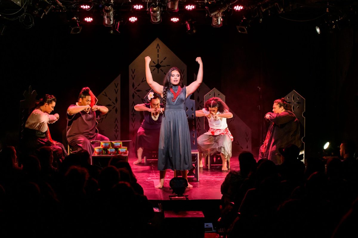 Wild Dogs Under My Skirt. Image is a group of Samoan women on a stage sitting on chairs in a semi circle and holding their fists together in front of them. In the middle stands another woman in a grey dress raising her arms triumphantly. The camera is at the back of the audience so some silhouetted heads are in the foreground.