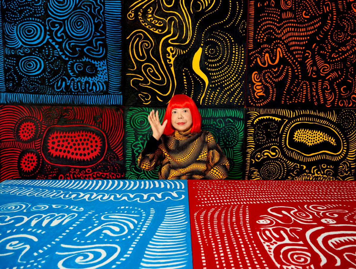 An elderly Japanese woman with bright red hair sits among multicoloured wall hangings and artworks.