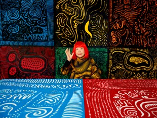 An elderly Japanese woman with bright red hair sits among multicoloured wall hangings and artworks.