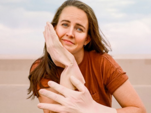 Zoë Coombs Marr. Image is a woman wearing oversized rubber gloves, outside with a beach behind her, leaning on her elbows at a table. She has long brown hair and is wearing a brown T shirt.