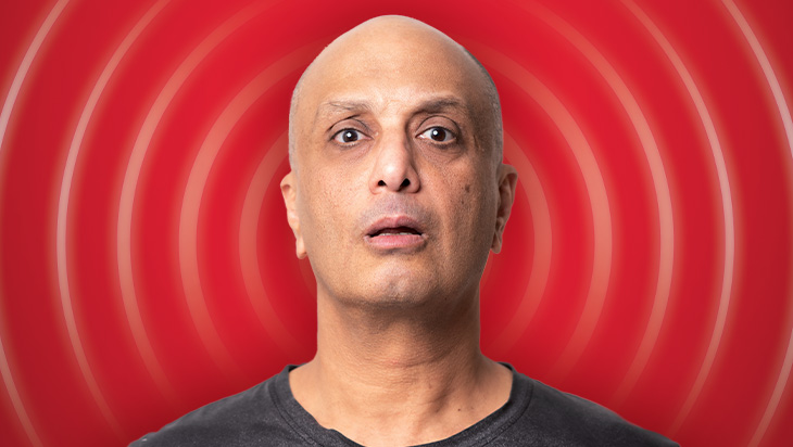 A headshot of a bald headed man of Egyptian heritage, wearing a black T shirt, against a red backdrop with curved lines emanating from his head to look as if he is broadcasting/radiating.