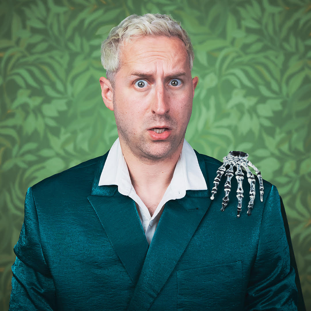 Andy Balloch. Image is a head and shoulders shot of a quizzical/concerned looking white man with bleached blond hair, a green jacket over a white open necked shirt and a plastic skeletal hand on his shoulder.