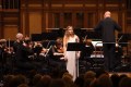 A white woman in a long sleeveless white dress stands in front of an orchestra singing, the conductor has his back to the camera. Sara Macliver, ASO, Grandeur.
