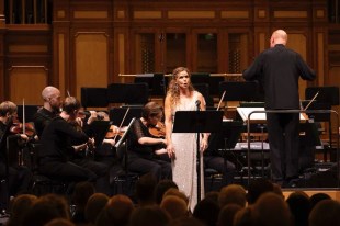 A white woman in a long sleeveless white dress stands in front of an orchestra singing, the conductor has his back to the camera. Sara Macliver, ASO, Grandeur.