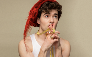 A young white man wears a white vest and a red Tudor hat with a long feature. He has his hands steepled in front of his face pensively.